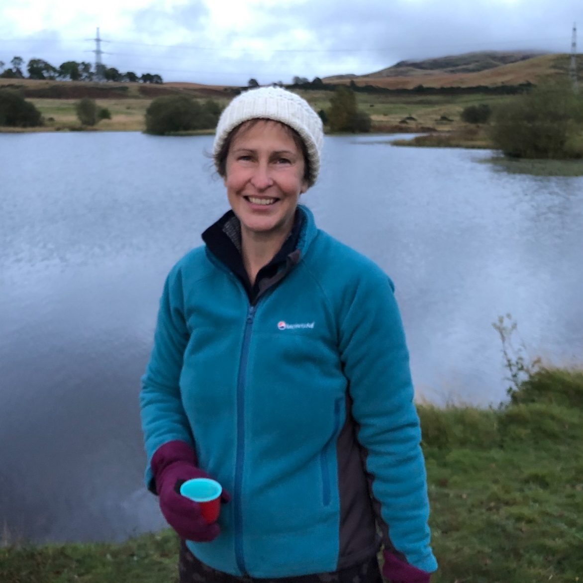 Woman in a blue jacket holding a cup, standing by a lake with hills in the background.